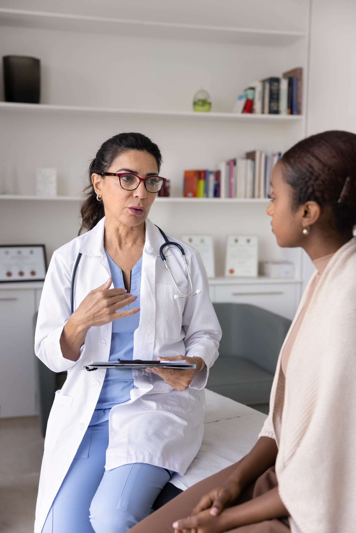 Serious middle aged Hispanic doctor woman giving consultation to young African patient sitting on couch in examination room, asking questions, holding file with medical records